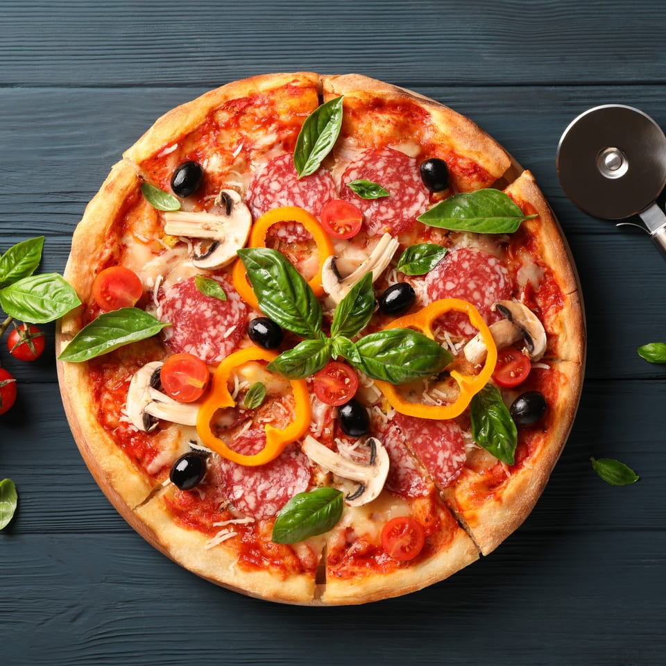 Guido's Pizza with pepperoni, mushrooms, bell peppers, olives, tomatoes, and basil on a dark wooden table.